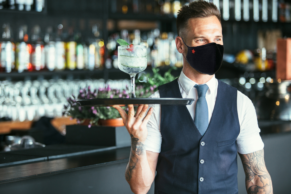 Een ober met een masker houdt een dienblad met een cocktail in een bar.