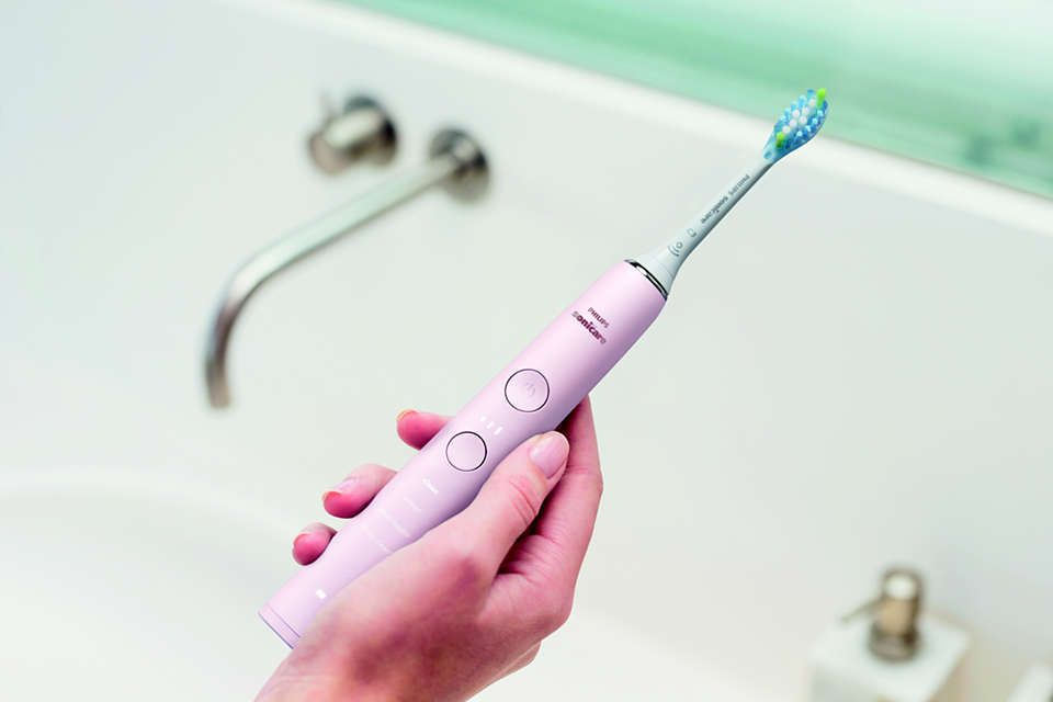 Pinke elektrische Zahnbürste, die von einer Hand gehalten wird. Die Zahnbürste steht neben einem Waschbecken.