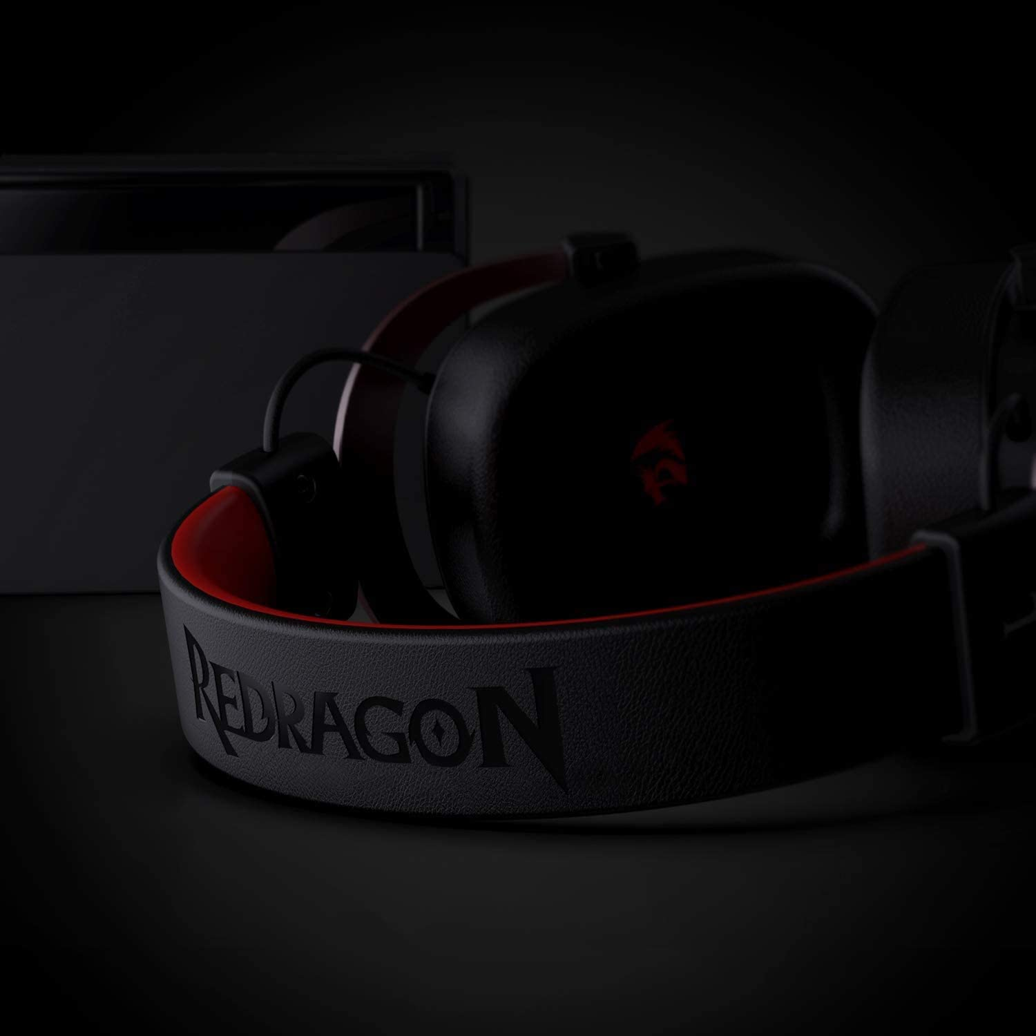 Fekete Redragon gaming headset sötét felületen, fekete játékkonzollal a háttérben.