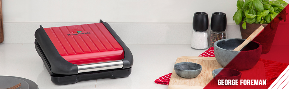 Piros George Foreman grill fekete díszítéssel. A háttérben vágódeszkák, tálak és bazsalikom látható.