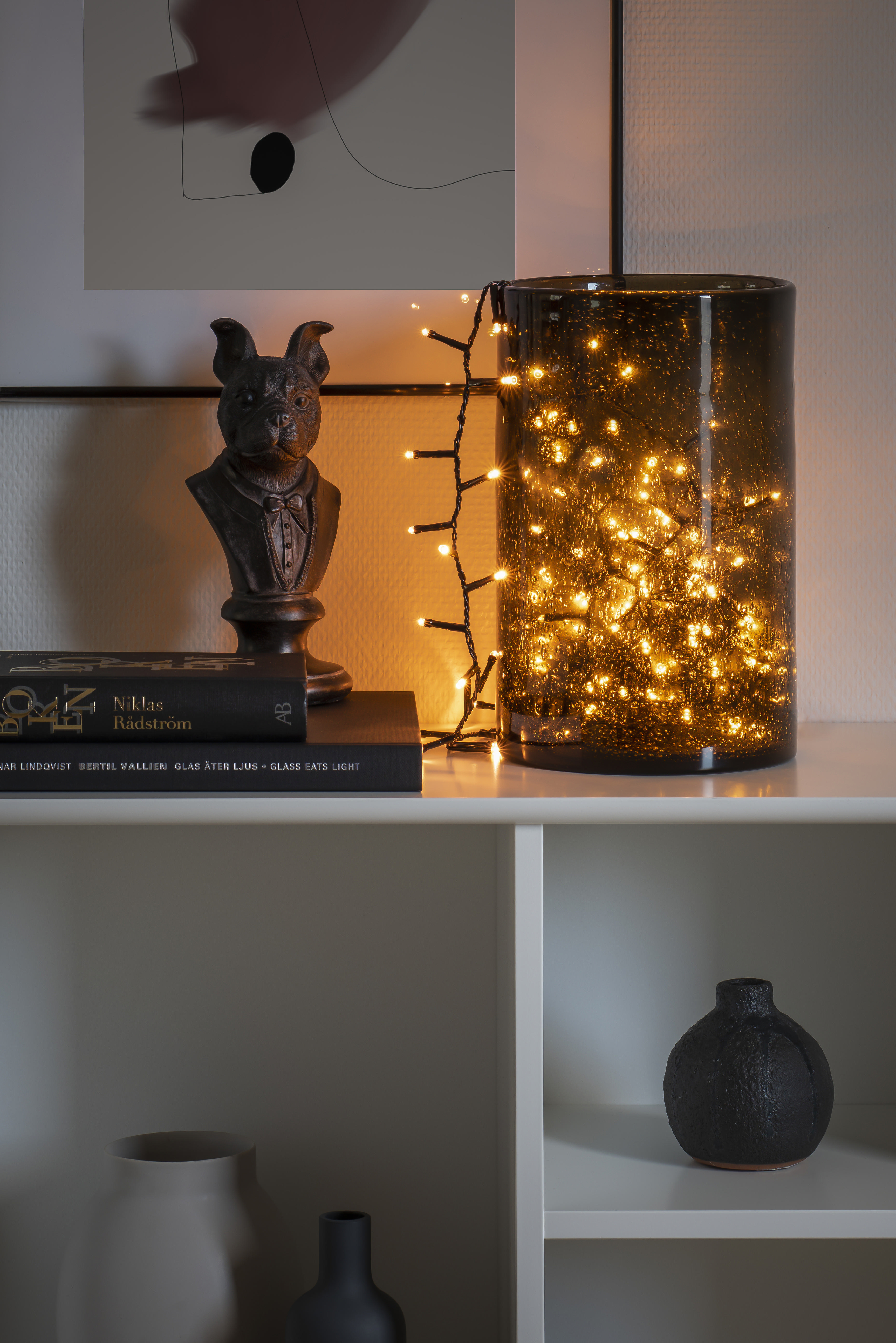 Dekorative Szene mit einer Lampe mit Lichtern darin, einer Statue, Büchern und einem Regal mit einer Vase.