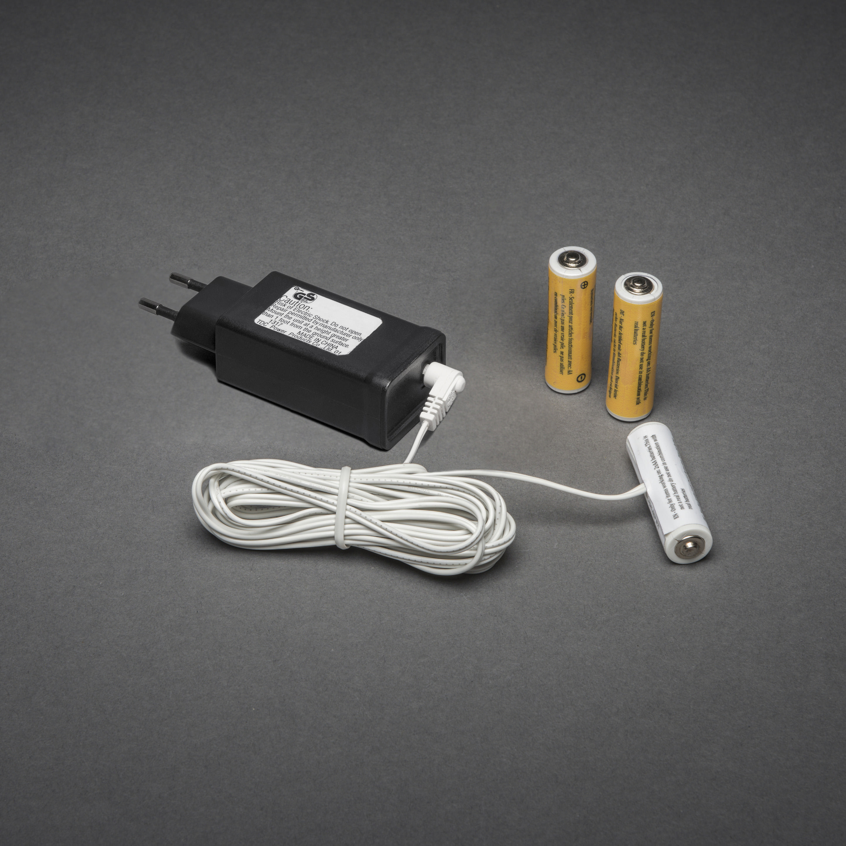 Ladeset: schwarzes Ladegerät, weißes Kabel und zwei gelbe Batterien. Auf grauer Oberfläche.