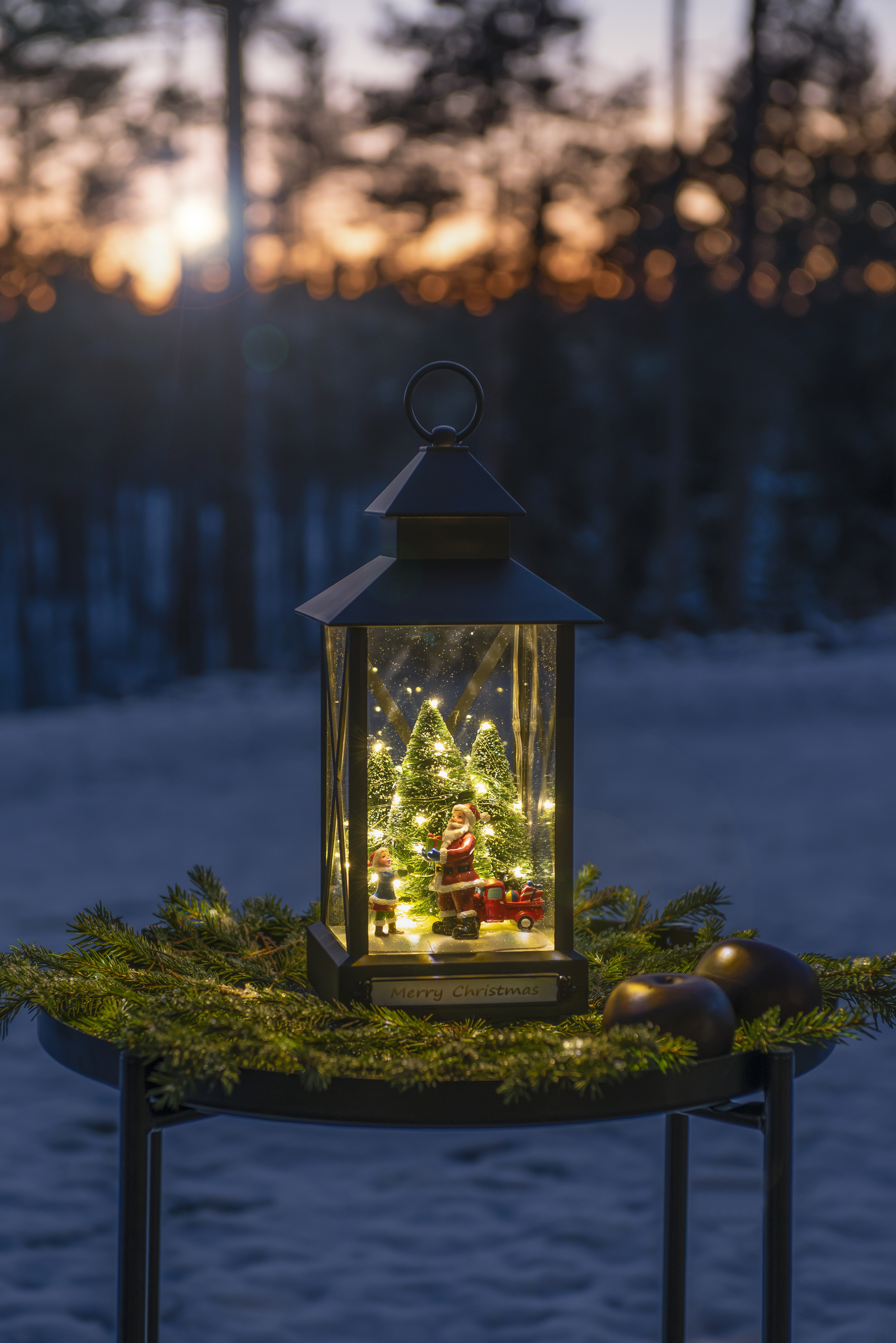 Eine Weihnachtslaterne mit Weihnachtsmann und Kind darin, auf einem Tisch mit Kiefern, vor einem verschwommenen Sonnenuntergang.