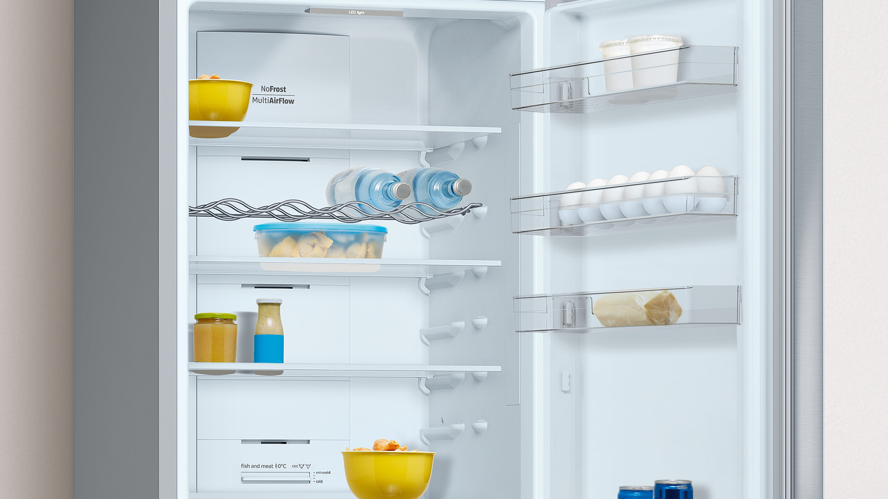 Interior de refrigerador abierto con estantes que contienen alimentos, incluyendo frutas, frascos y botellas de agua.