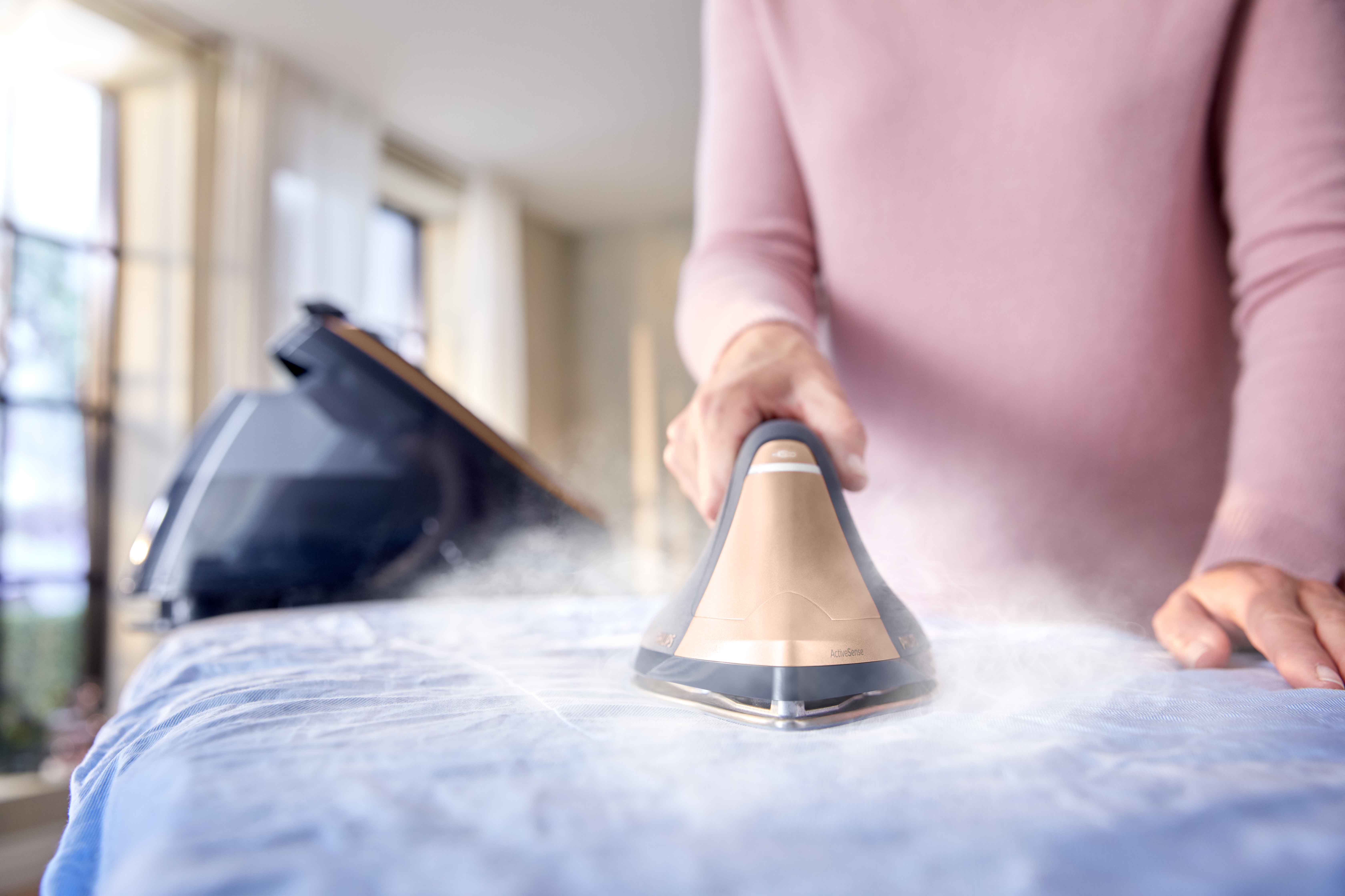 Bir kişi, ütü masasında, buharlı ütü ile mavi bir gömlek ütülüyor.