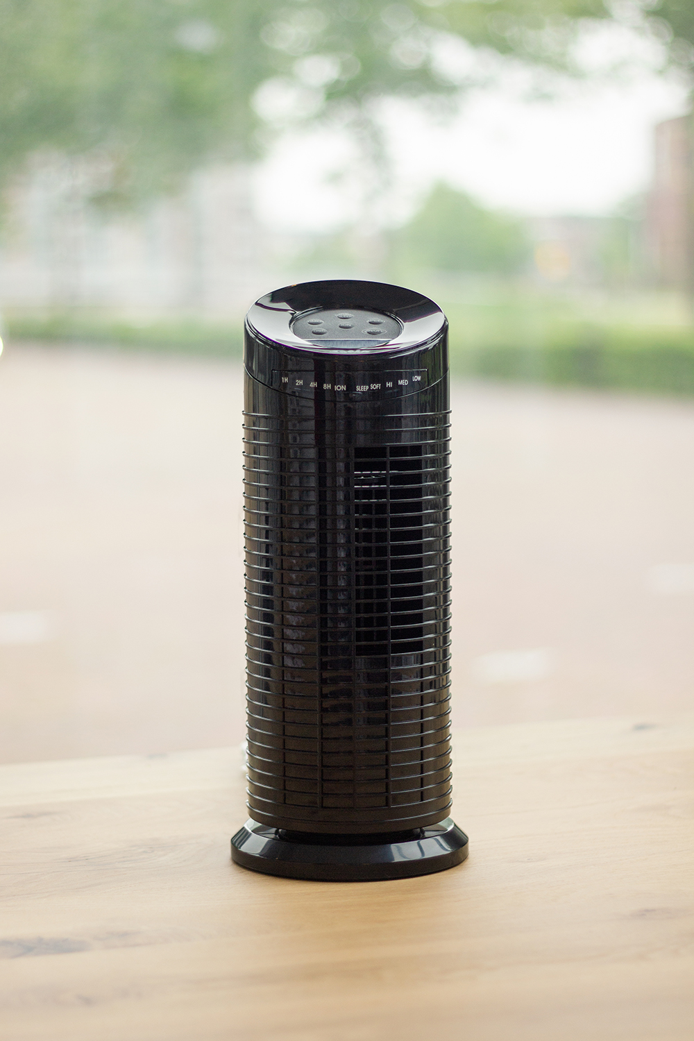 Un purificateur d'air noir est posé sur une table en bois. L'arrière-plan montre une fenêtre floue avec du feuillage vert et un bâtiment.