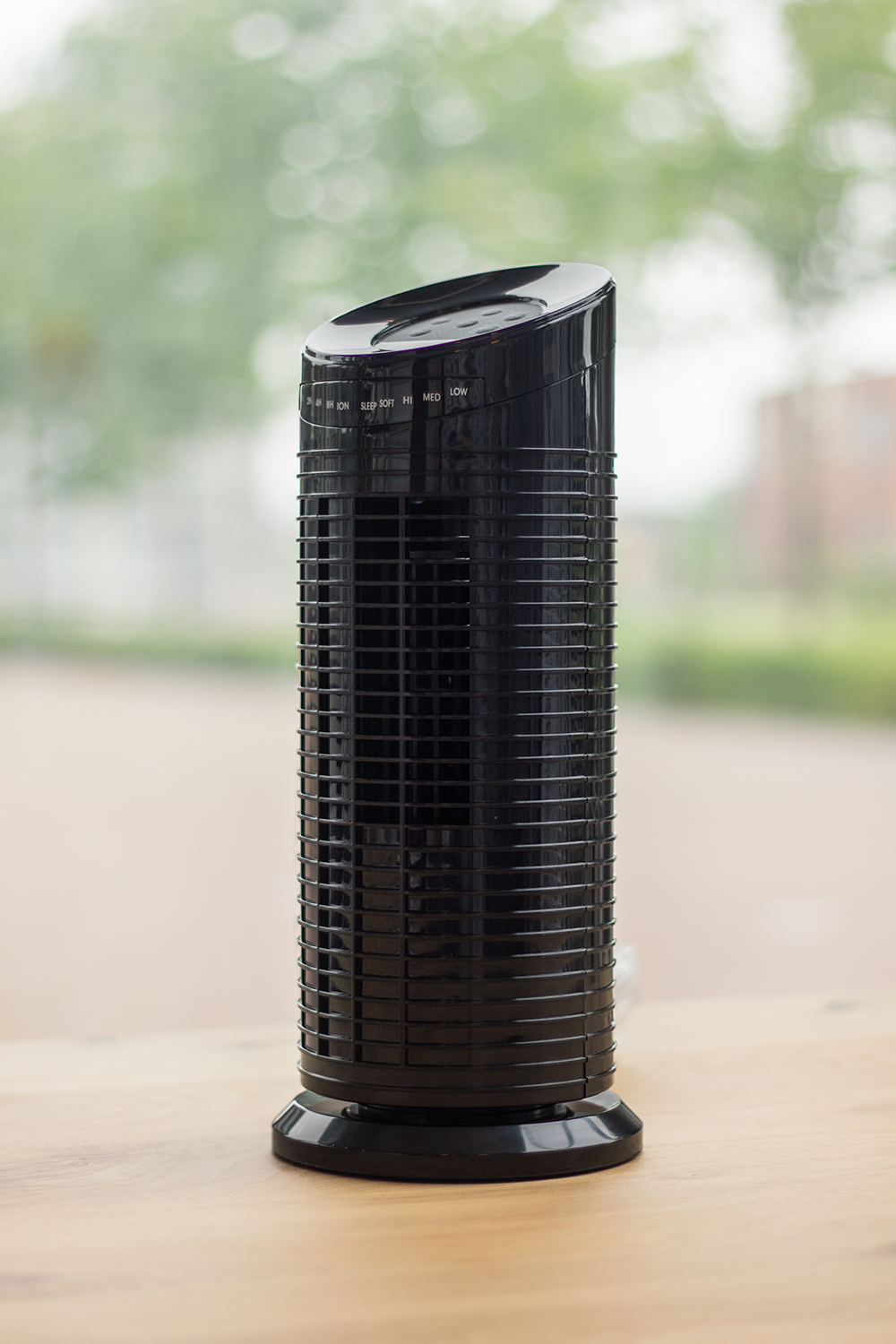 Un purificateur d'air noir est posé sur une table en bois. Il a un design cylindrique avec des boutons de commande sur le dessus. Une fenêtre est en arrière-plan.