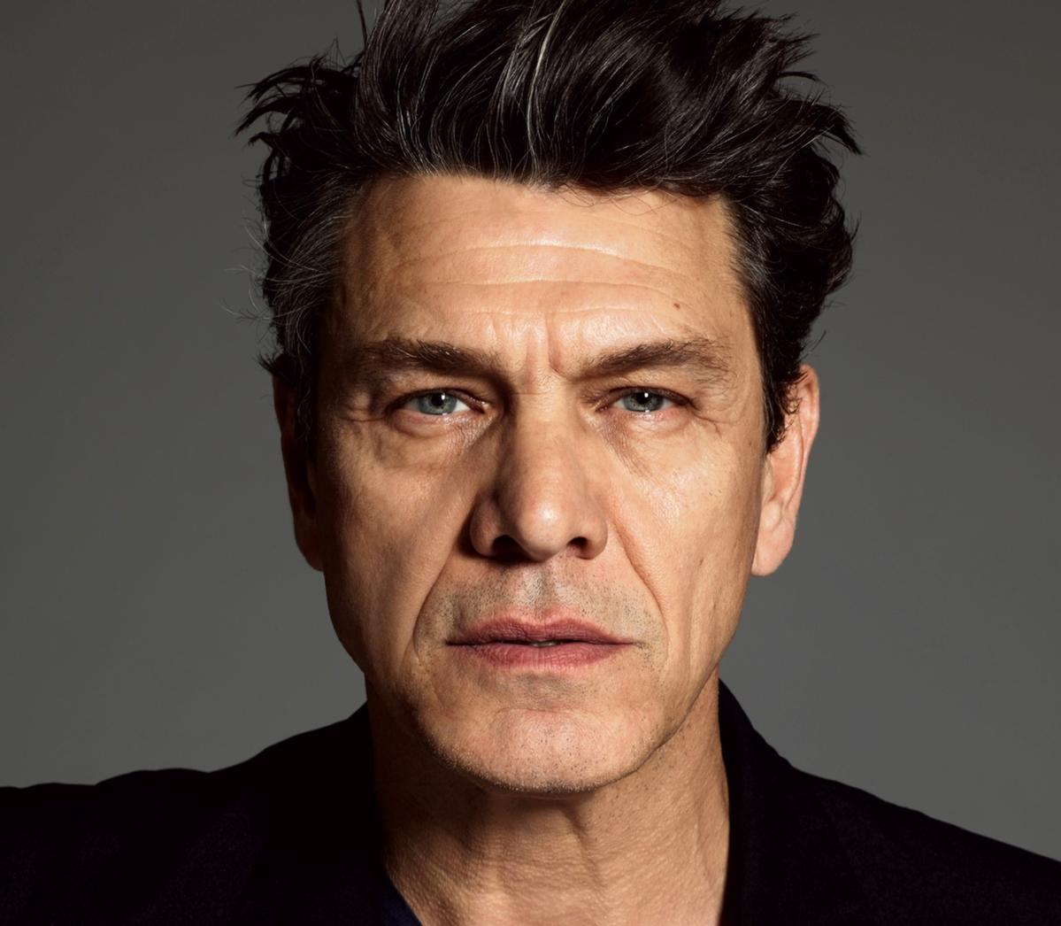 Gros plan sur le visage d'un homme. Cheveux gris, yeux bleus, veste sombre. Fond gris.