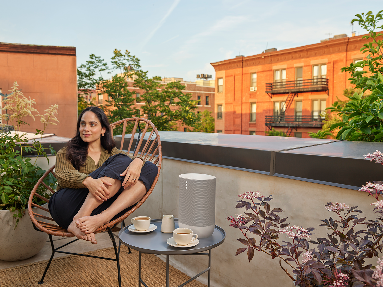 Vrouw op een terras, zittend in een stoel met een luidspreker en koffie. Bakstenen gebouwen op de achtergrond.