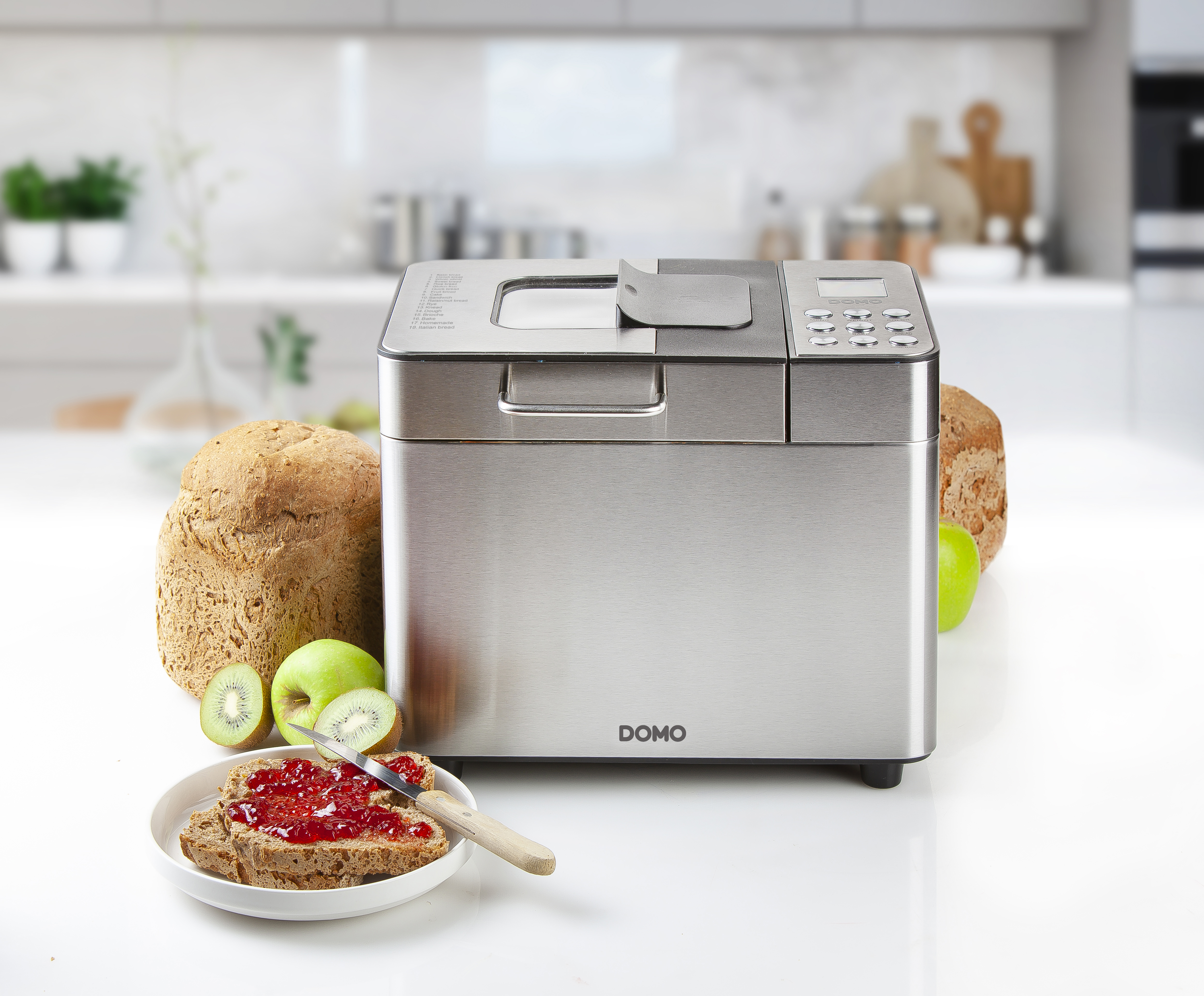 Une machine à pain argentée avec du pain et des fruits sur une surface blanche.