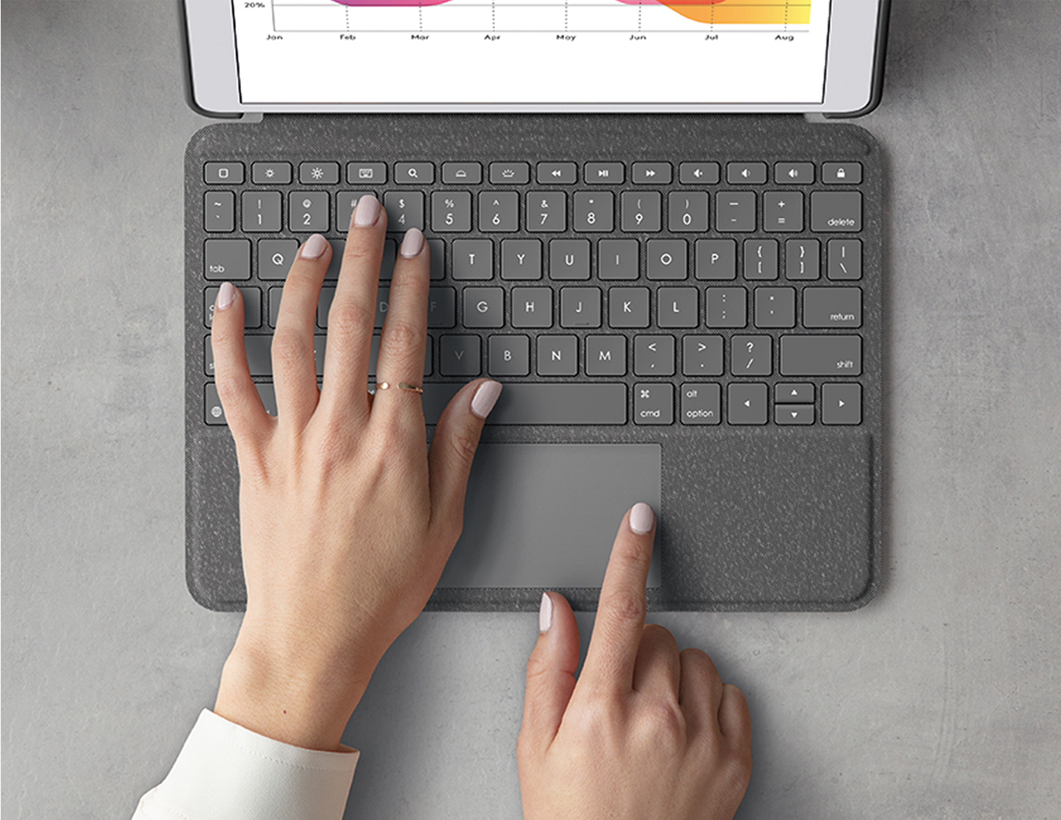 Hands on a gray keyboard with a trackpad, a tablet screen is barely visible.