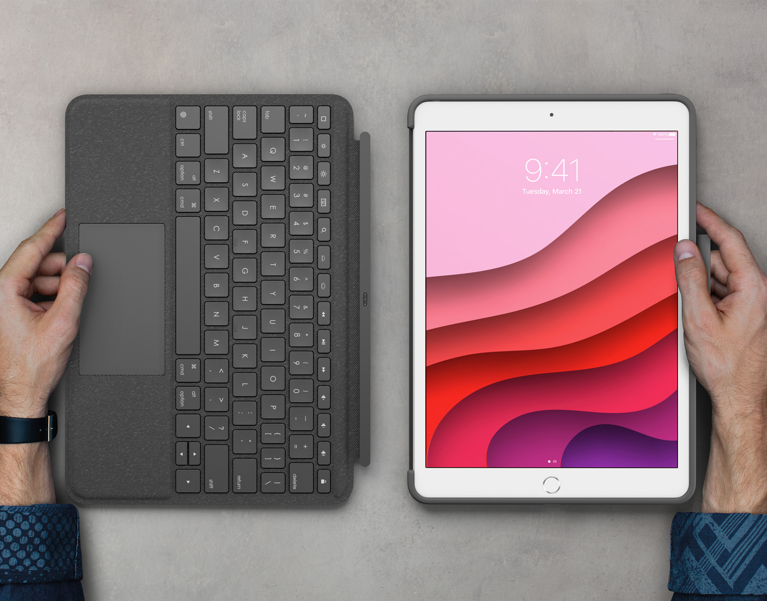 Hands holding a keyboard and iPad. Keyboard is dark grey, iPad has a pink and red layered screen.