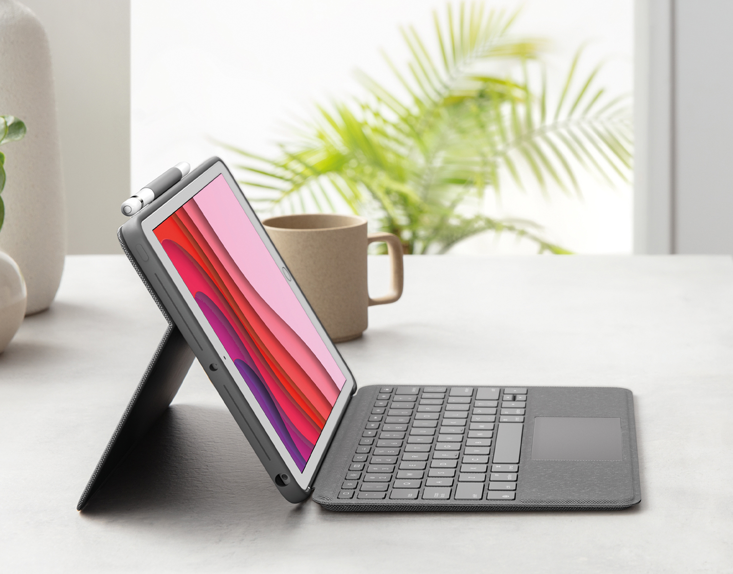 Tablet with keyboard case and stylus on a table, with window and plants in background.