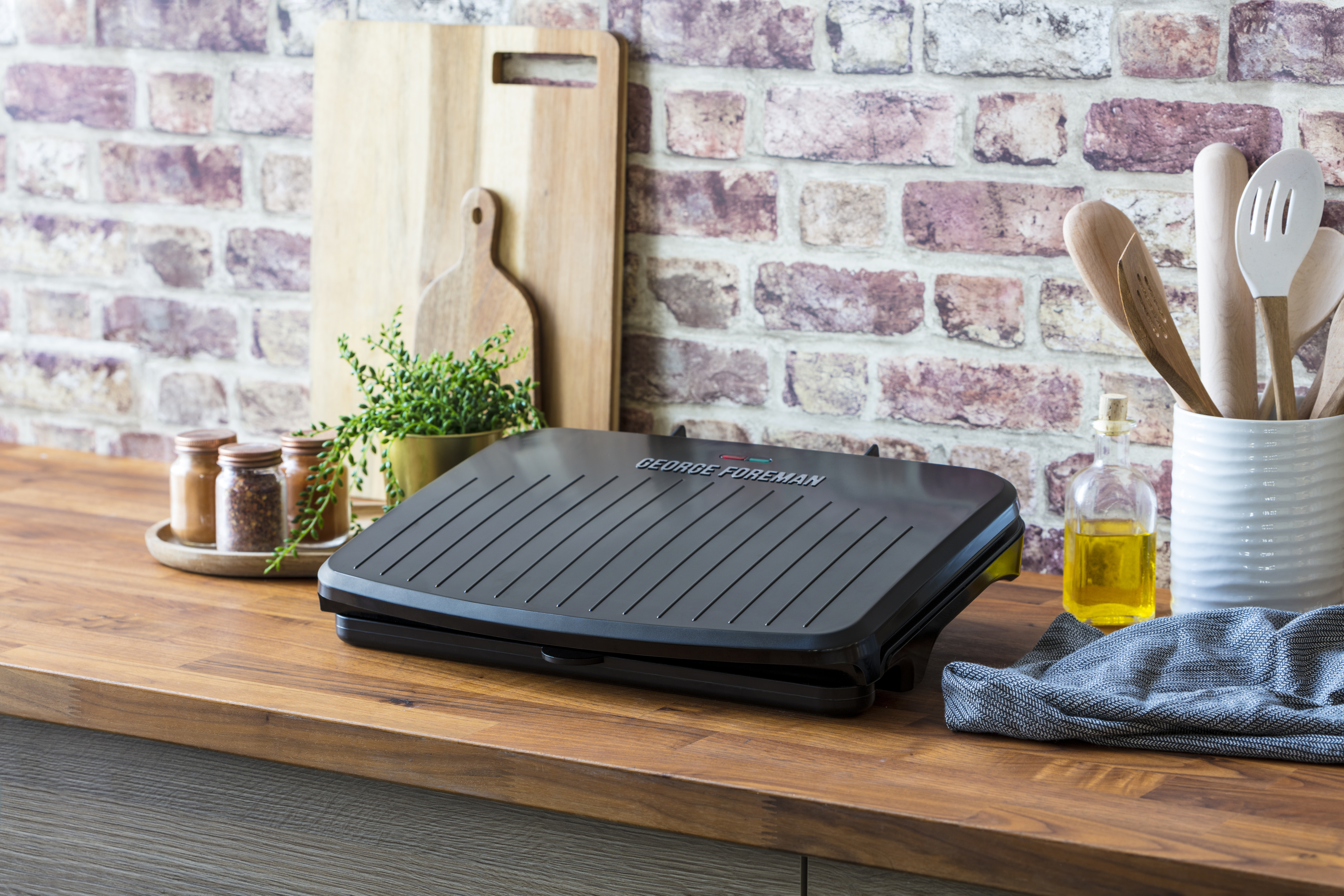 Egy fekete George Foreman grill fa pulton, téglafallal a háttérben, edényekkel.