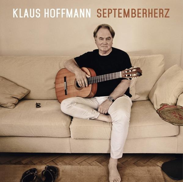 Klaus Hoffmann sitzt auf einem Sofa, hält eine Gitarre, trägt ein schwarzes Hemd und eine weiße Hose.