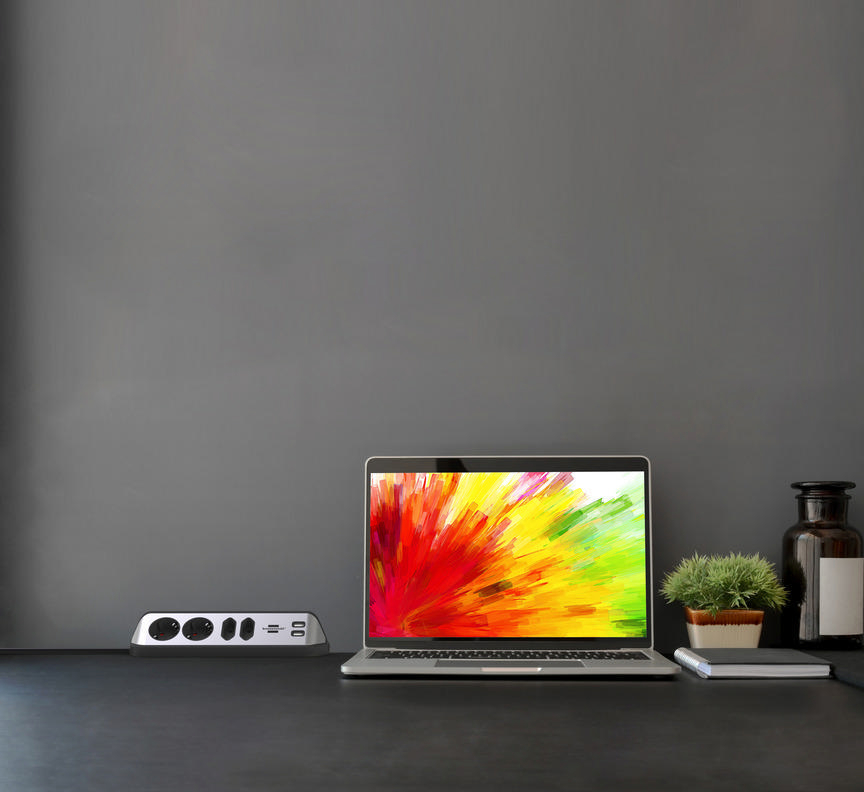 Laptop mit farbenfrohem Display, Steckdosenleiste, Pflanze und einem Glas auf einem schwarzen Schreibtisch.