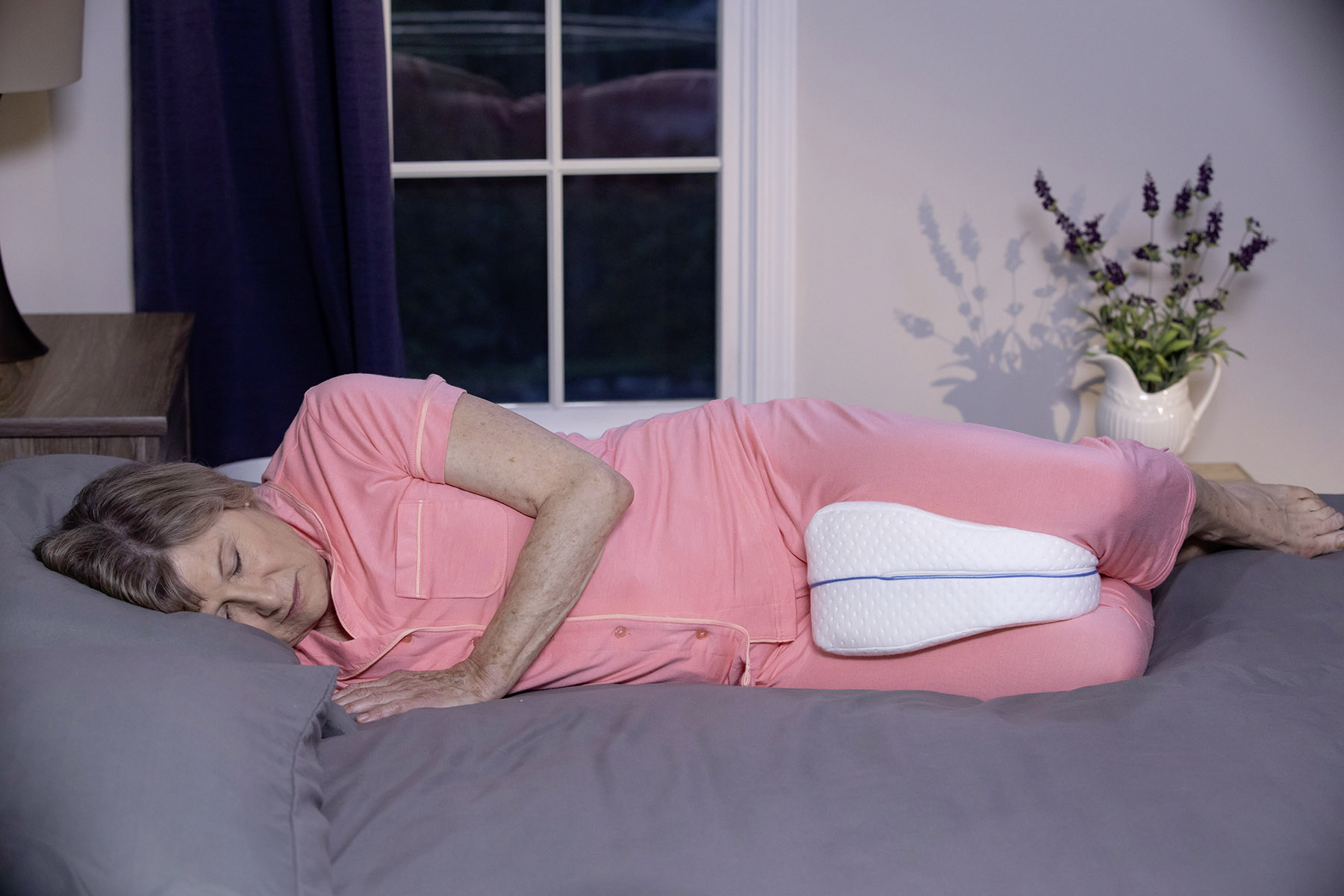 Eine ältere Frau schläft in rosa Pyjamas auf der Seite, ein Kissen zwischen den Knien. Im Hintergrund ein Fenster, Vorhang und Blumen.