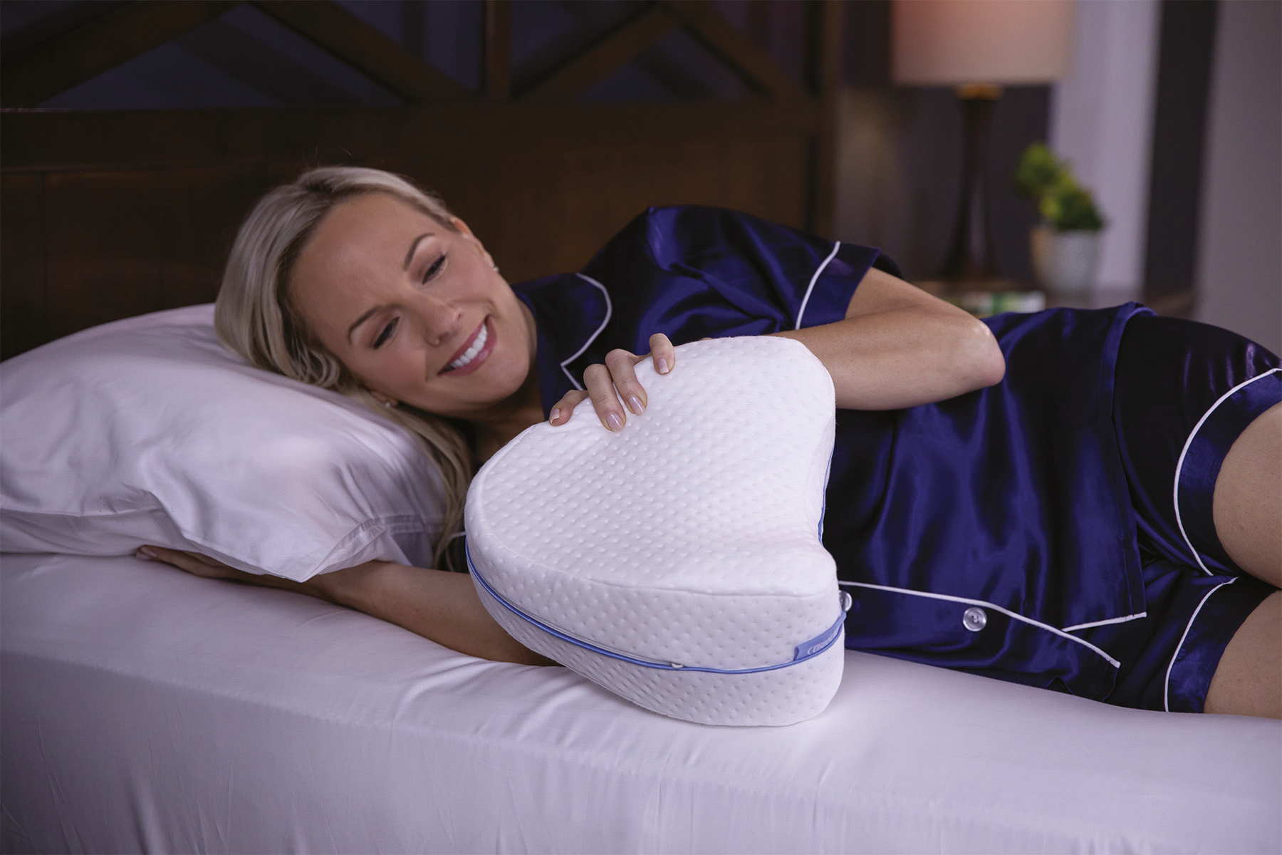 Eine Frau in blauem Pyjama lächelt, während sie auf einem Bett mit einem weißen Kissen und einem Kniekissen liegt.