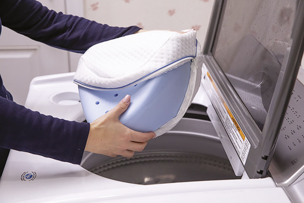 Eine Person legt ein blau-weißes Kissen in eine Waschmaschine. Der Deckel der Waschmaschine ist offen. Die Person trägt ein dunkelblaues Hemd.