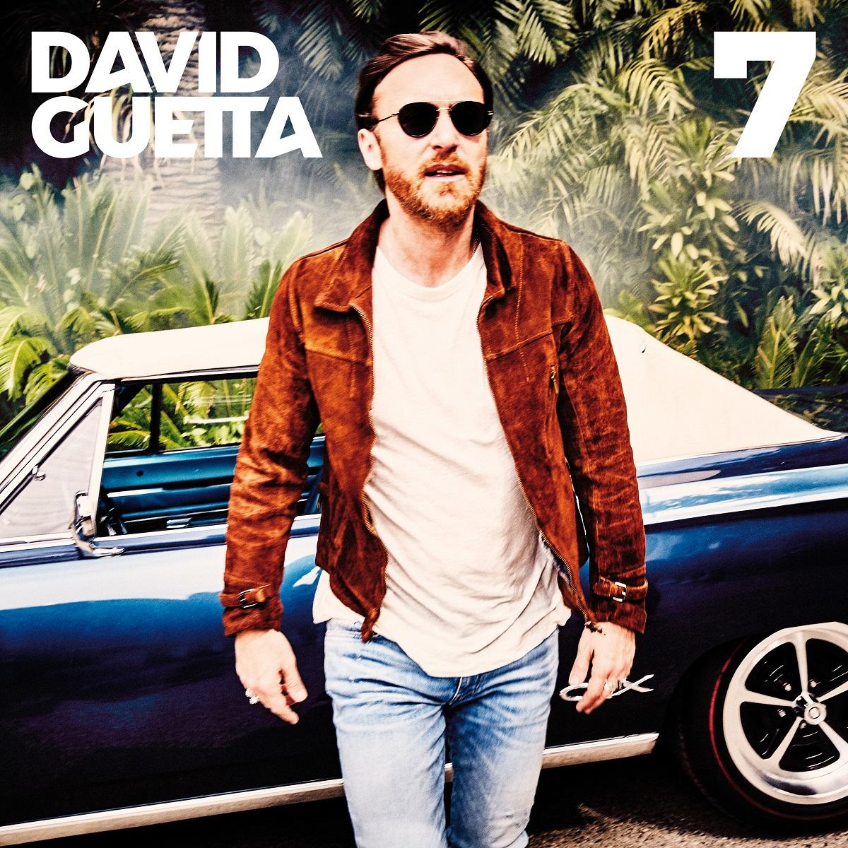 David Guetta napszemüvegben és barna dzsekiben egy autó mellett áll egy napsütéses napon.
