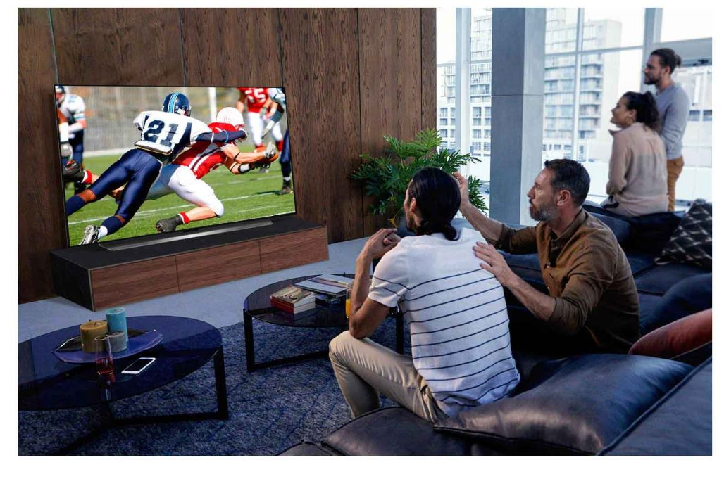 İnsanlar televizyonda Amerikan futbolu izliyor. İki adam ön planda. Diğerleri arka planda. TV, ahşap bir stand üzerinde.