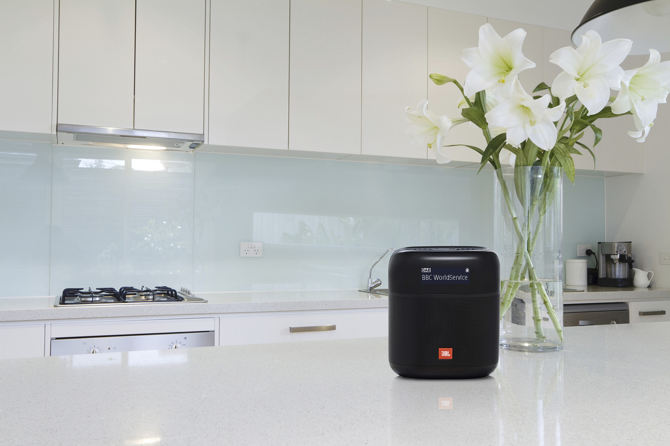 A black JBL speaker on a white countertop near white lilies.