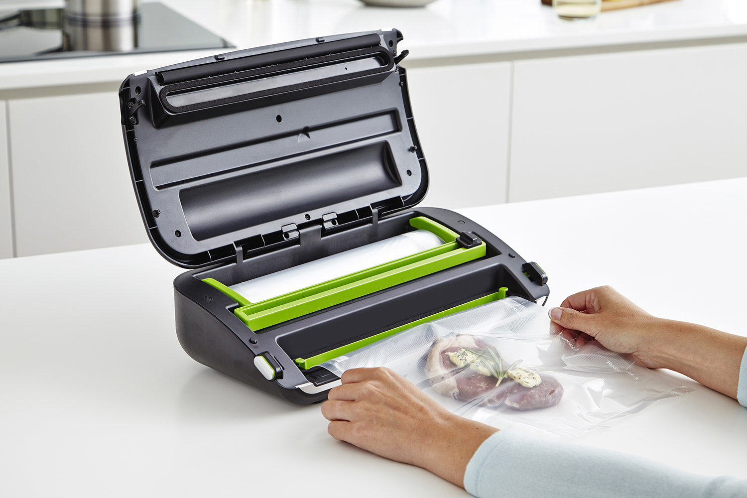 Vacuum sealer with hands sealing a bag containing meat. Black and green machine, white surface.