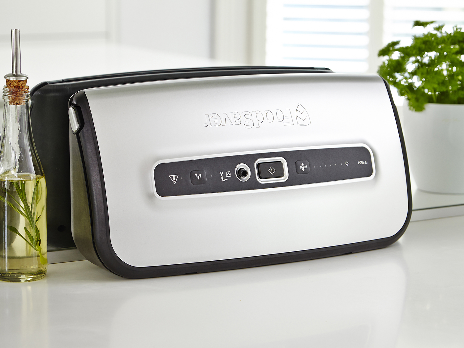 A silver FoodSaver vacuum sealer with a bottle of oil and a plant.
