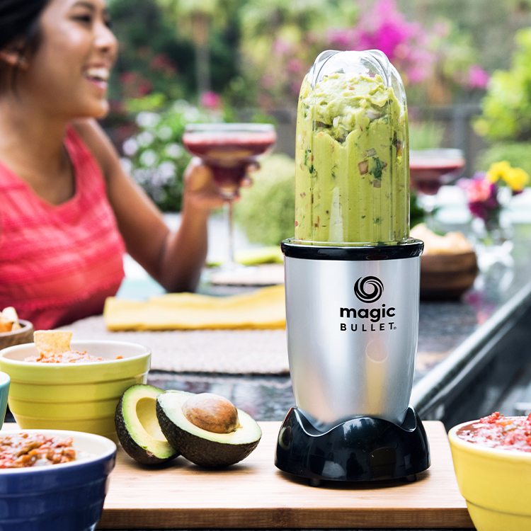 Person with a Magic Bullet blender making guacamole outdoors, with ingredients on a table.
