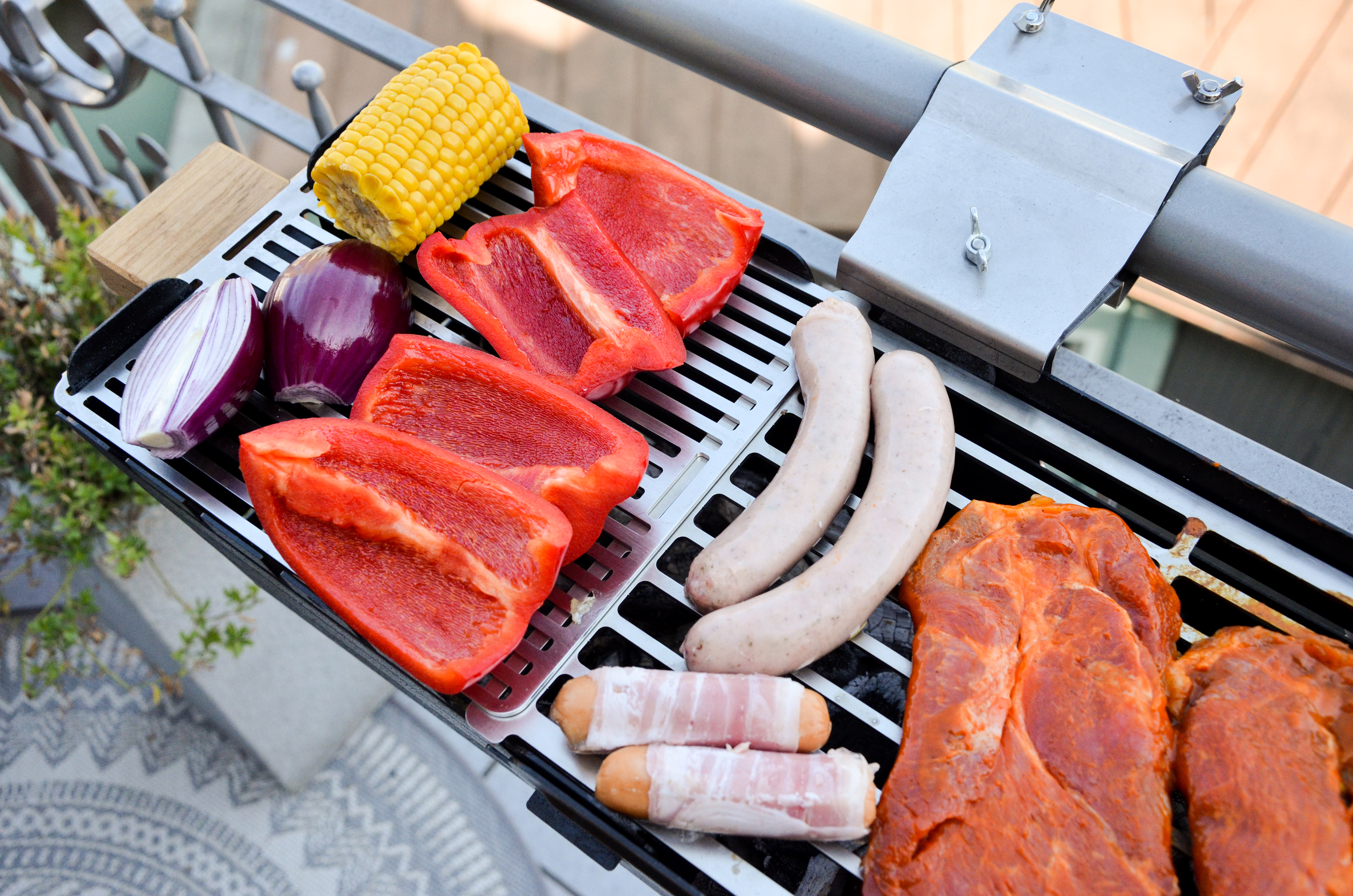 Ein Grill mit verschiedenen Lebensmitteln: Mais, rote Paprika, Würstchen und mariniertes Schweinefleisch. Auf einem Balkongeländer.