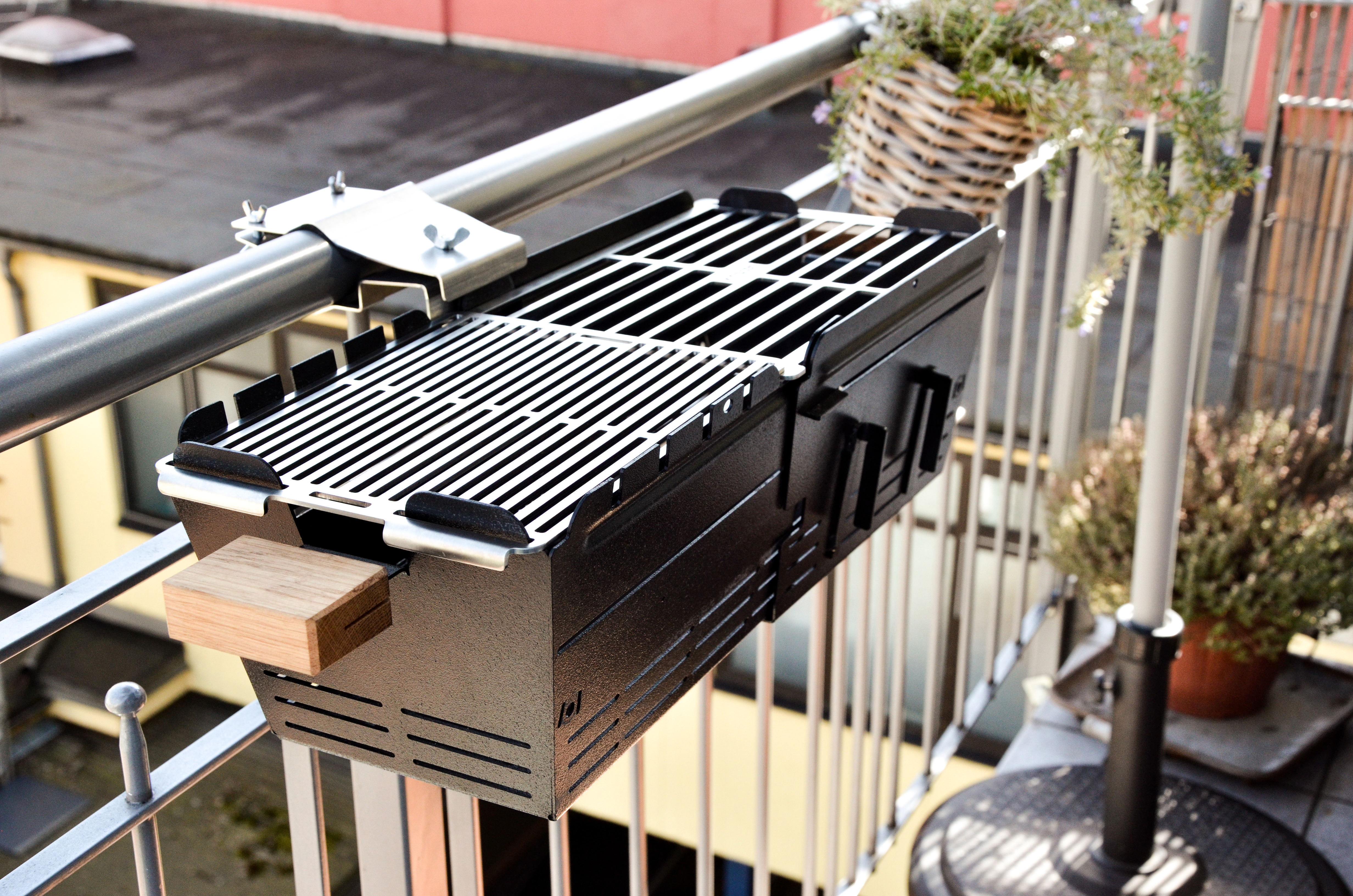 Schwarzer Grill auf Balkon. Hängt über dem Geländer. Pflanzen und Sonnenschirm im Hintergrund.