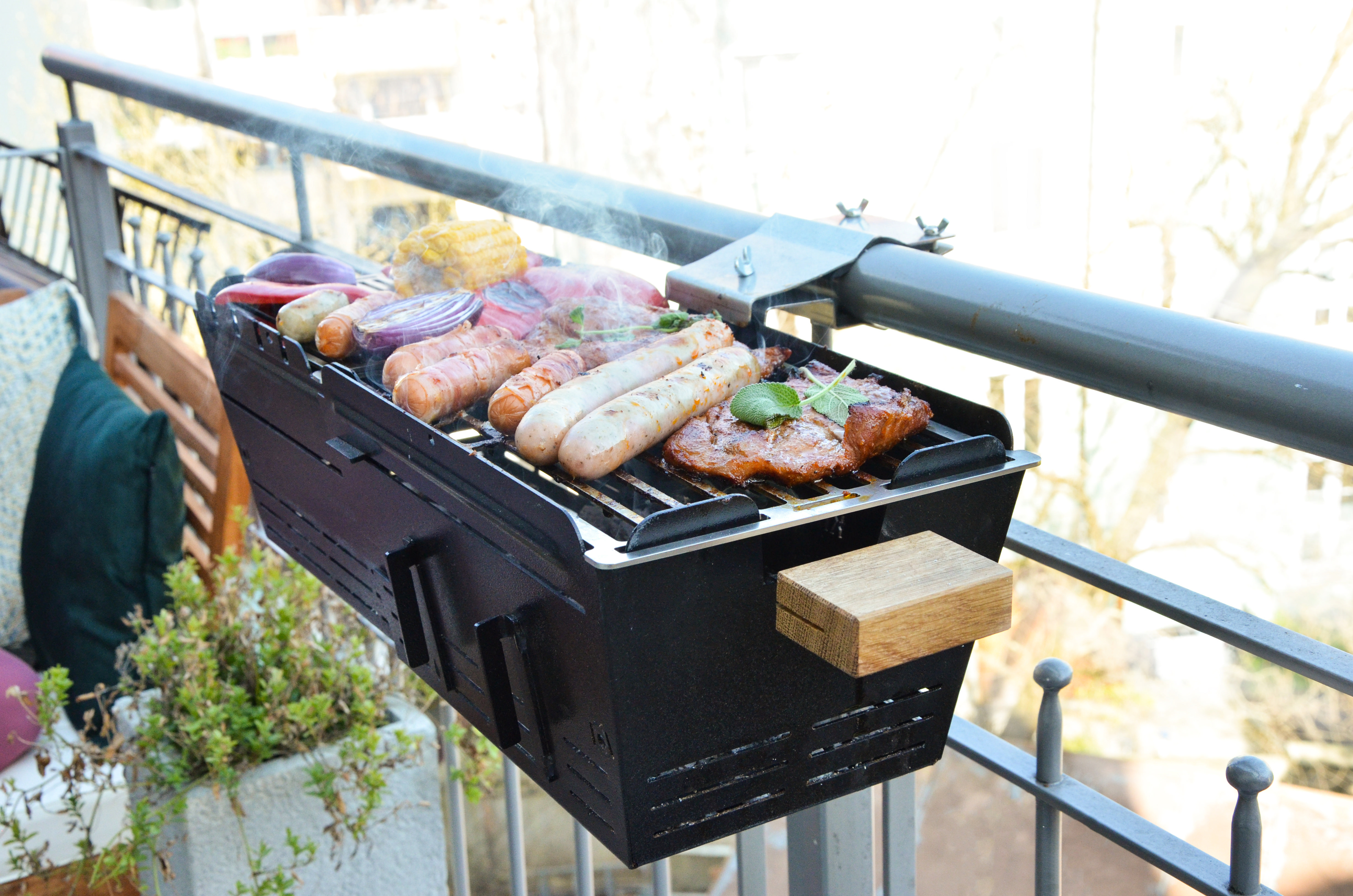 Balkongrill mit grillendem Essen. Enthält Würstchen, Mais und Gemüse.