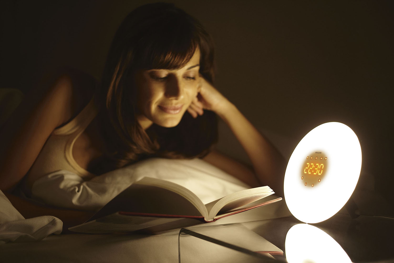Een vrouw leest een boek in bed met een Philips-wekker die de tijd aangeeft.