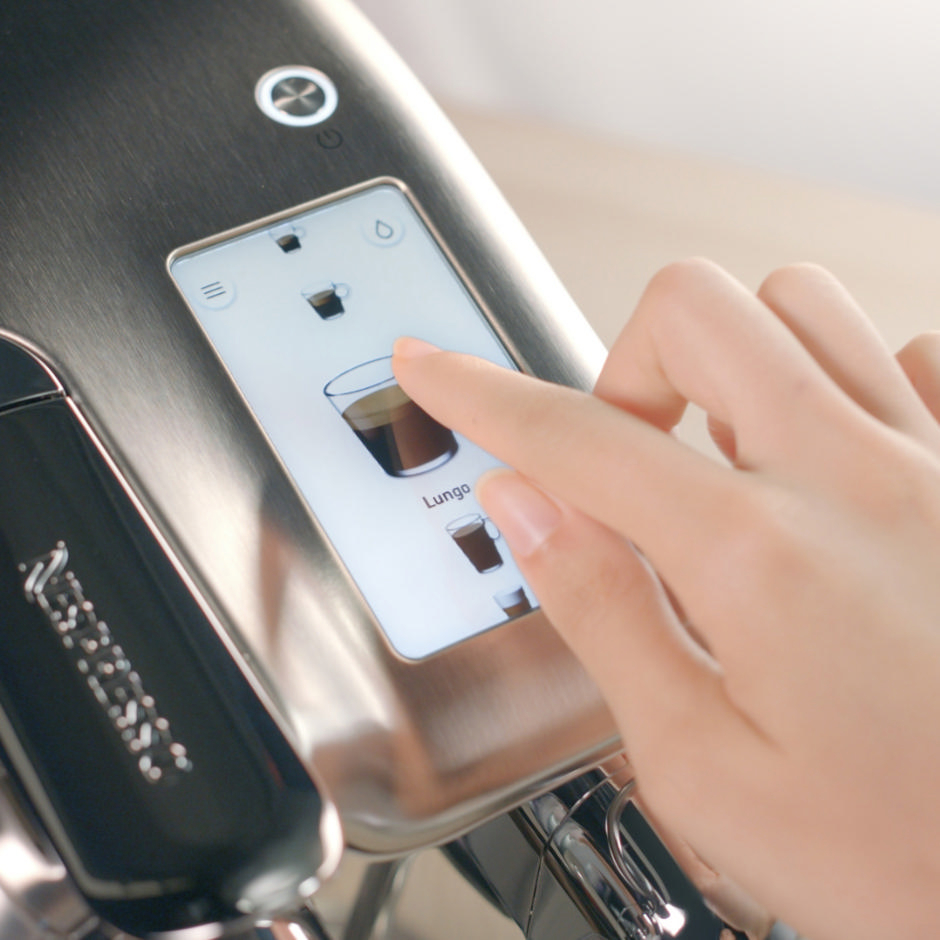 Eine Hand berührt einen Kaffeeautomatenbildschirm. Der Bildschirm zeigt ein Kaffeebild und die Beschriftung 'Lungo'.