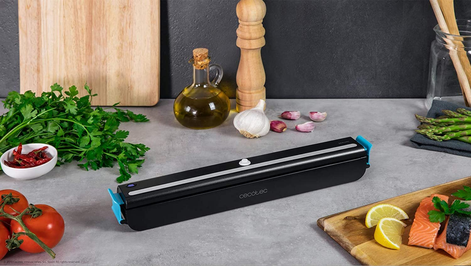 Escena de cocina con envasadora al vacío, comida y utensilios. Encimera gris. Tablas de cortar de madera.