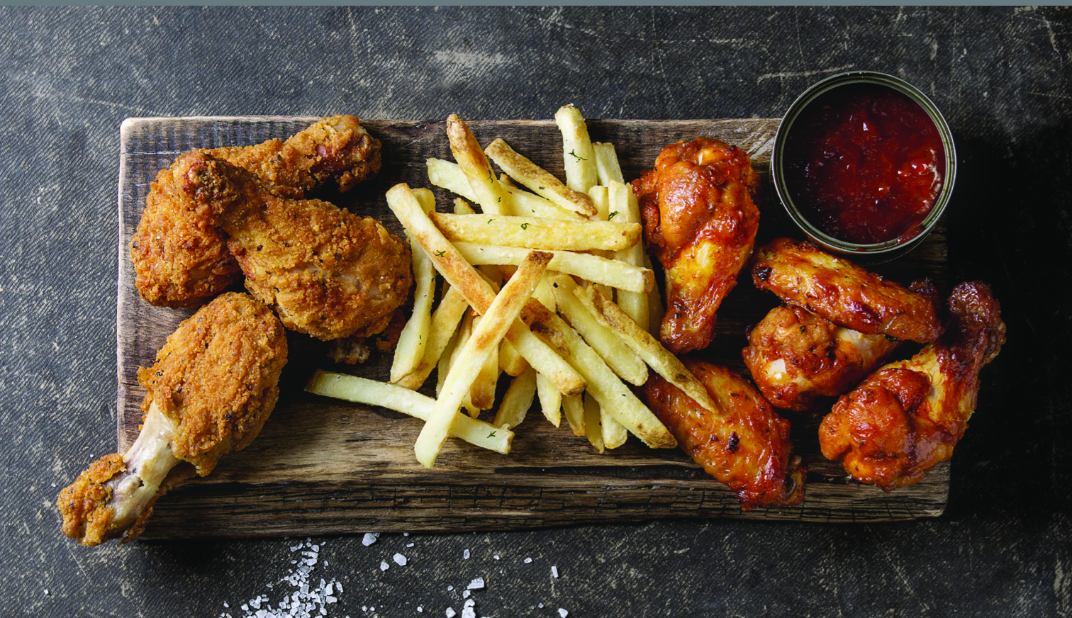 Wooden board with fried chicken, fries, and sauce. Dark background with salt.