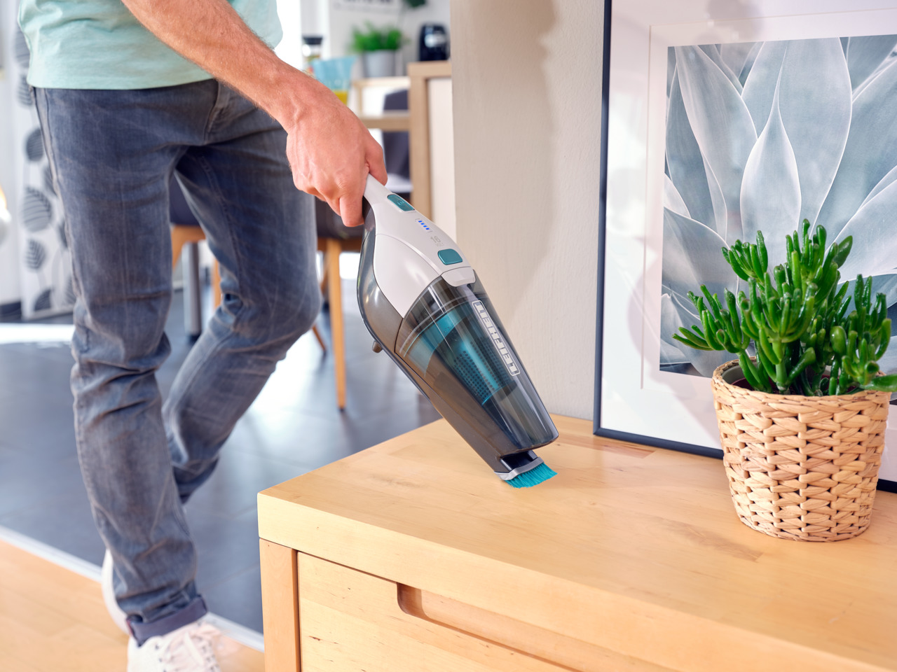 Person saugt Holztisch mit Handstaubsauger. Pflanze, Bilderrahmen sichtbar.