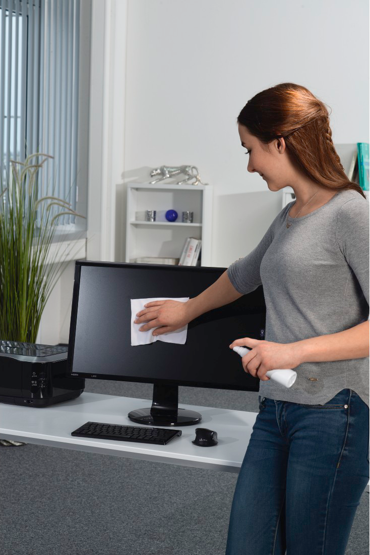 Eine Frau reinigt einen Computermonitor mit einer Sprühflasche und einem Tuch.