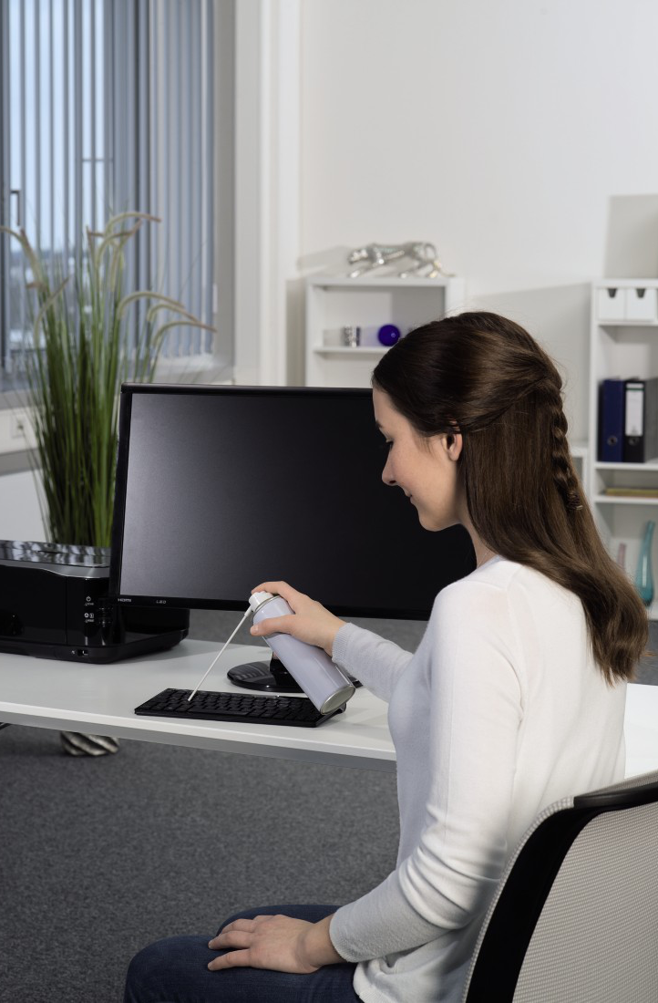 Frau sprüht Tastatur mit Reiniger. Büro mit Monitor und Pflanzen.