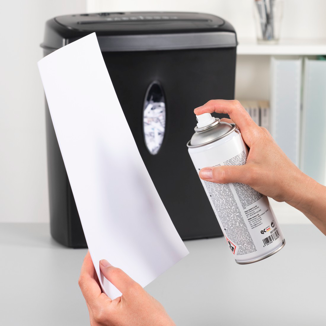 Eine Person besprüht ein weißes Blatt Papier mit einer Dose. Im Hintergrund befindet sich ein schwarzer Aktenvernichter.