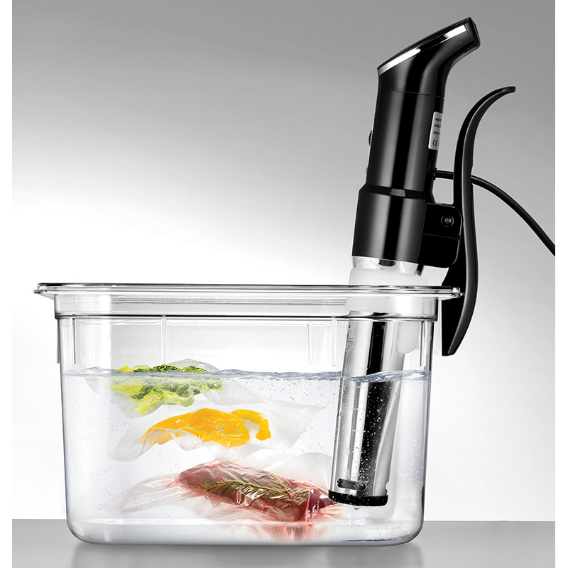 Sous-Vide-Kochaufbau. Schwarzes Gerät in Wasser mit Essen in Beuteln. Klarer Behälter.