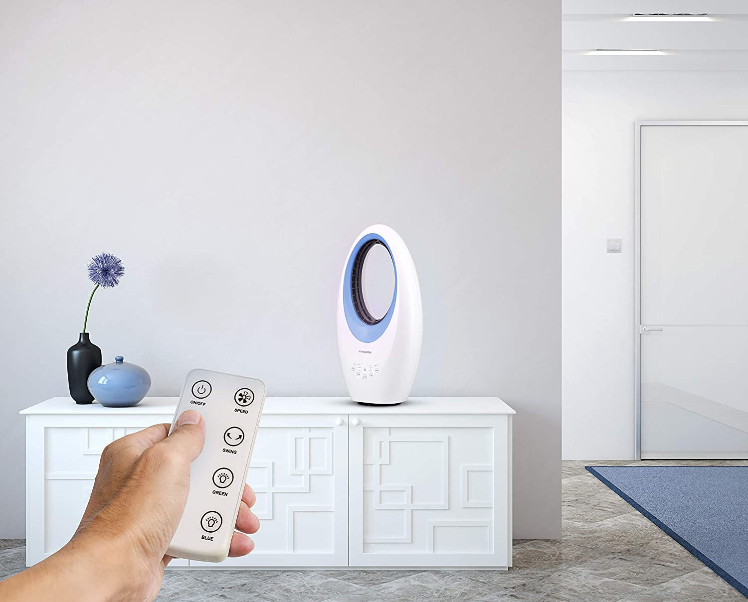 Egy kéz távirányítót tart, ventilátor egy fehér szekrényen egy szobában.