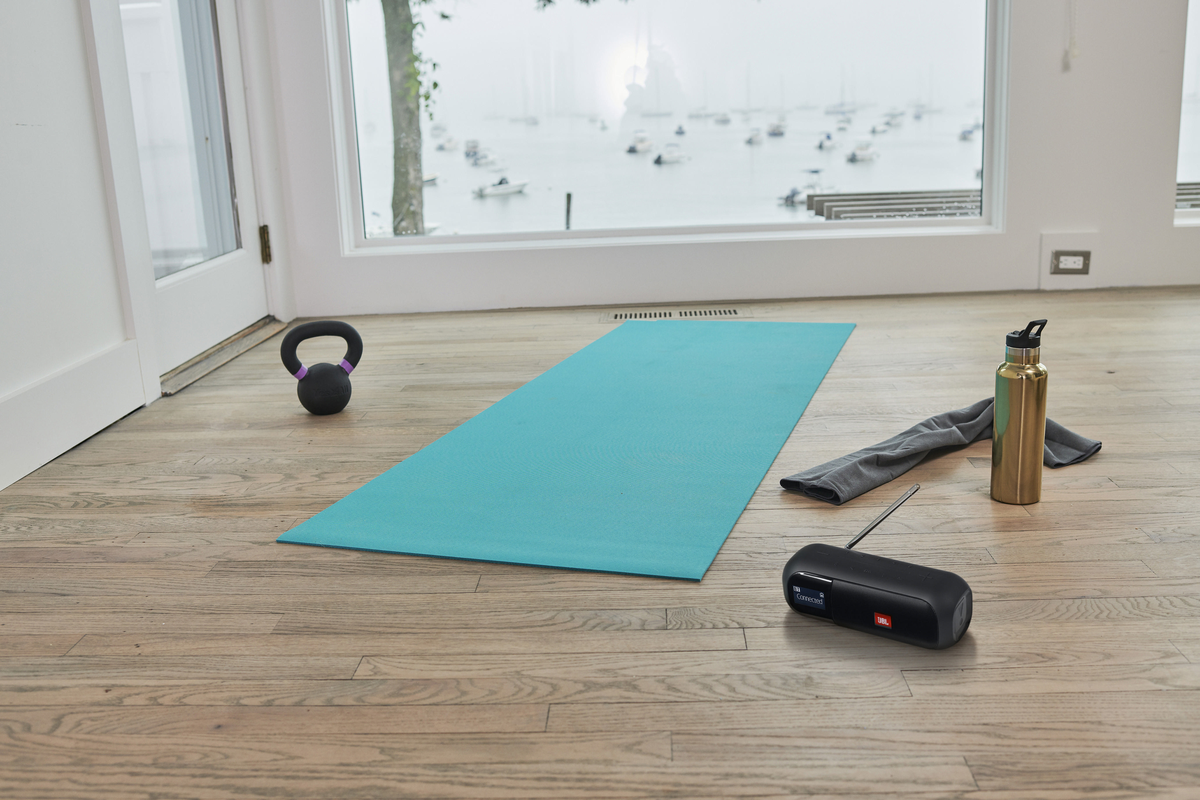 Eine Yogamatte, eine Kettlebell, ein Handtuch, eine Wasserflasche und ein Lautsprecher liegen auf einem Holzboden.