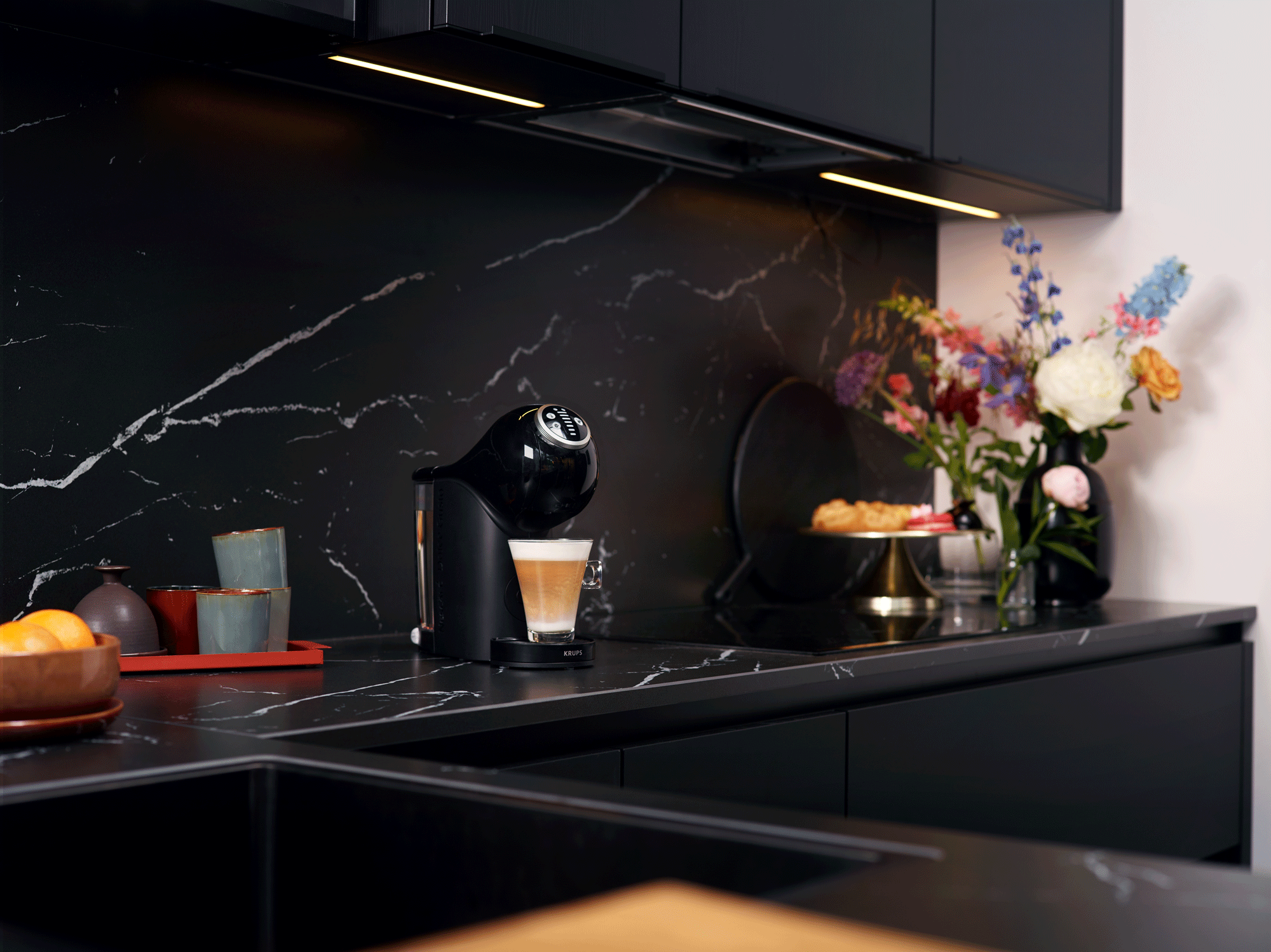 Keuken met zwart aanrecht, koffiezetapparaat, bloemen en gebak.