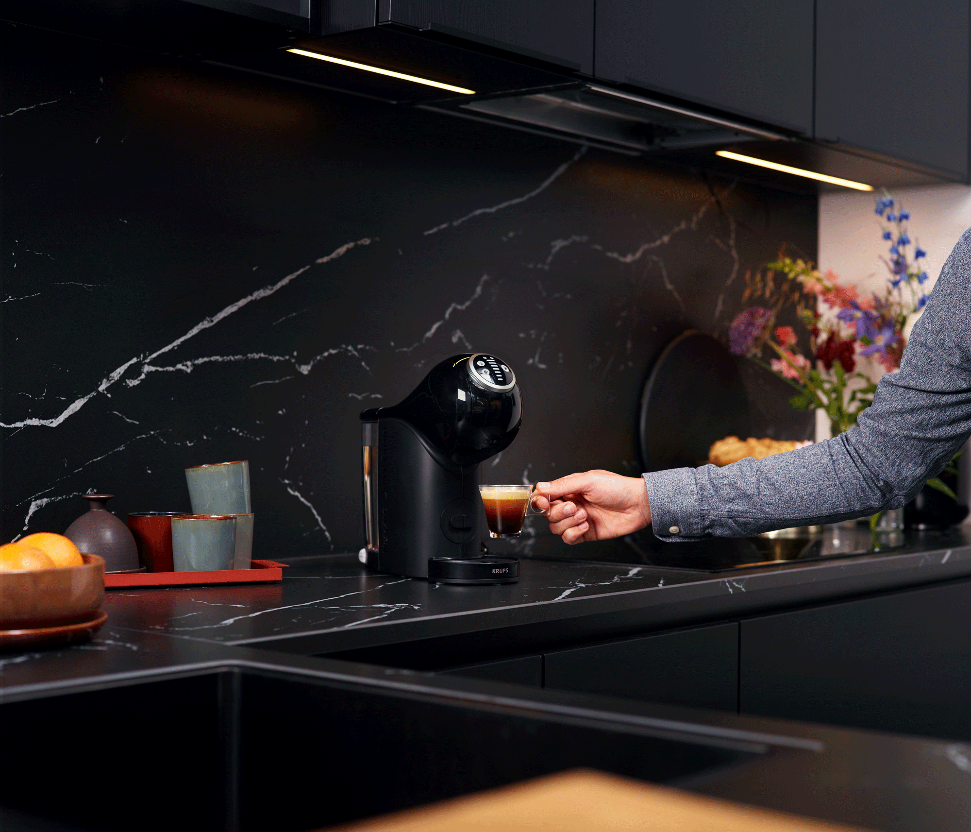 Een hand reikt naar een kopje koffie op een zwart aanrecht, met een koffiemachine.