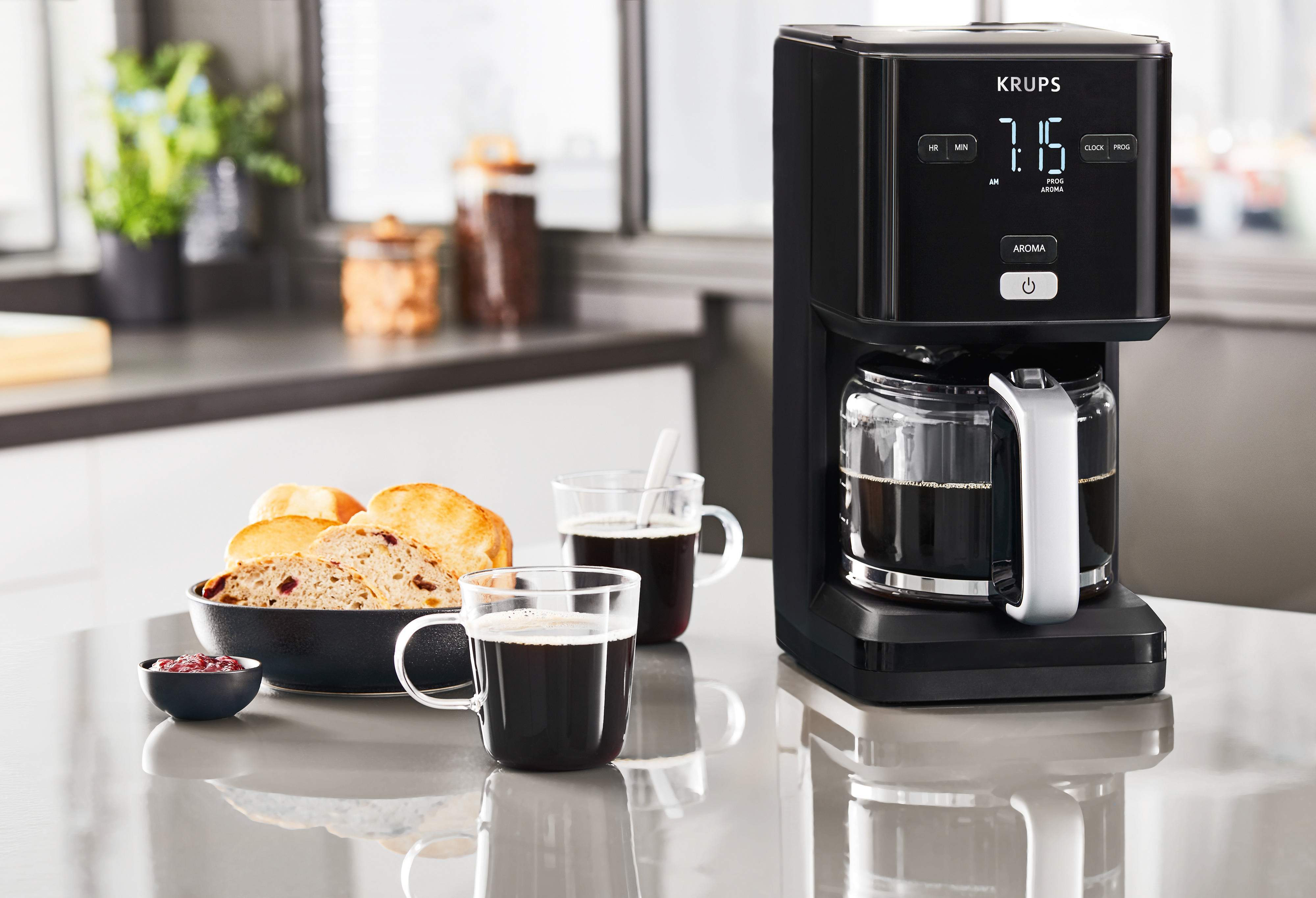 Eine schwarze Krups Kaffeemaschine mit Glaskaraffe, zwei Tassen Kaffee, Brot und Marmelade auf der Theke.