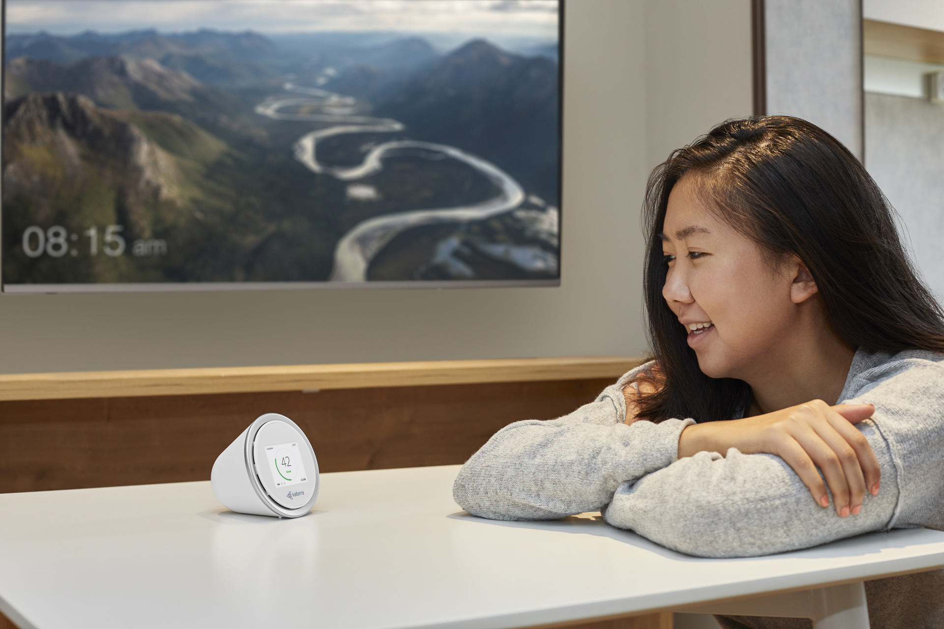 Frau lächelt eine weiße Smart-Home-Gerät auf einem Tisch an. Fernseher im Hintergrund.