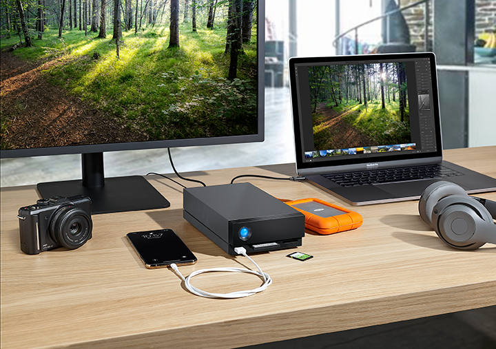 Ein Schreibtisch-Setup mit Monitor, Laptop, Kamera, Telefon, externer Festplatte und Kopfhörern. Ein Wald ist auf dem Bildschirm.