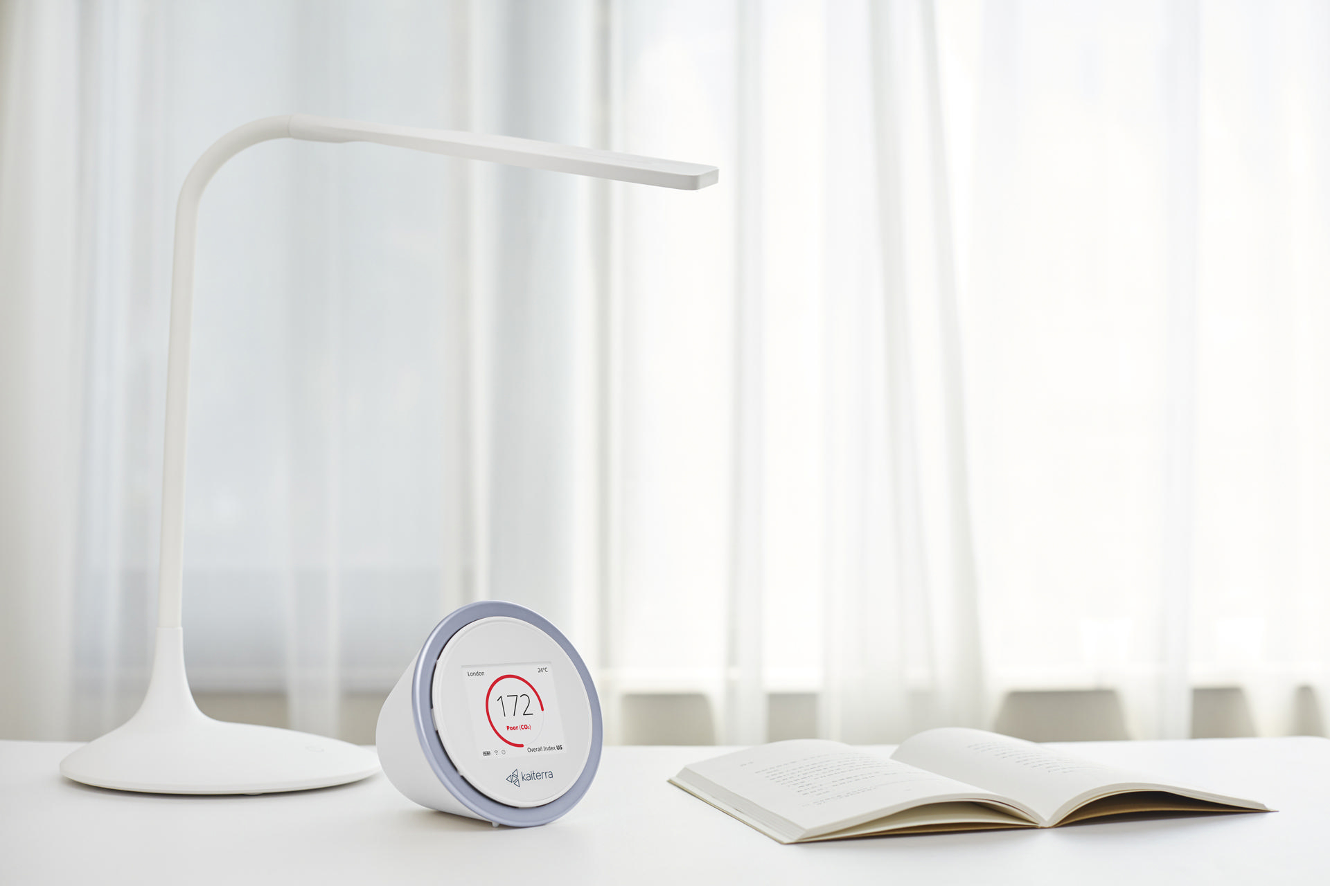 Weiße Schreibtischlampe, digitales Gerät und offenes Buch auf weißer Oberfläche.