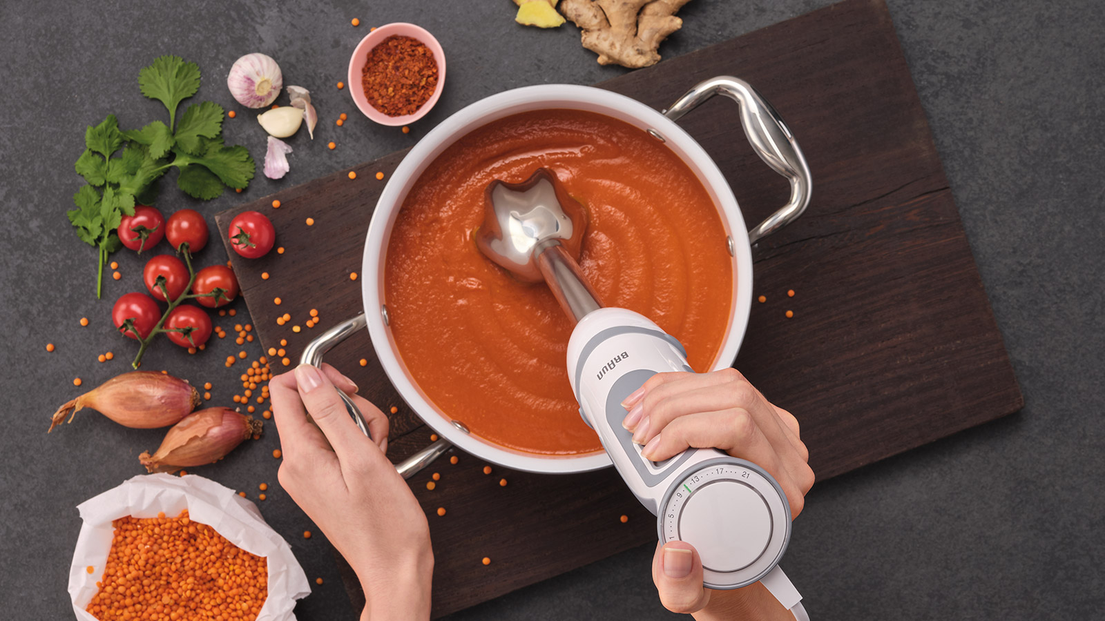 A person's hands using a stick blender in a pot of orange soup, with ingredients like tomatoes.
